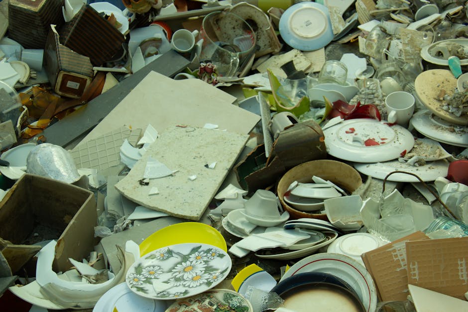 A large pile of broken and discarded household items, including shattered ceramic plates with floral patterns, cracked glassware, and miscellaneous kitchenware, is spread across a concrete pavement in