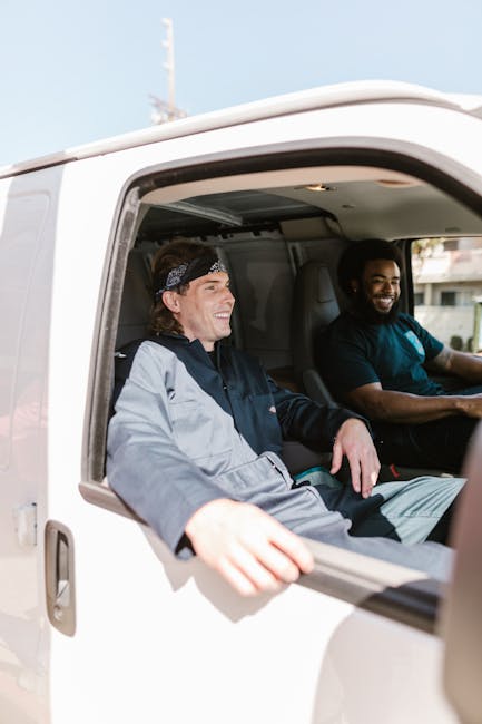 Two men are seated inside a white moving van, with the passenger-side window open, during a house relocation process. The man on the left, wearing a grey and black jacket and a dark headband, is smiling and looks relaxed. The man on the right, dressed in a dark T-shirt, is also smiling and appears to be the driver. Inside the van, visible behind them, are packing materials such as cardboard boxes, plastic wrapping, and fabric covers, along with some furniture, possibly chairs or small tables, prepared for transport. The scene is set outdoors, with natural daylight illuminating the interior of the van and parts of a residential area visible in the background. This image captures the loading process during furniture transport, characteristic of professional removal services like those offered by Man with Van Cranham, as part of packing and moving activities associated with home relocation within the Cranham area.