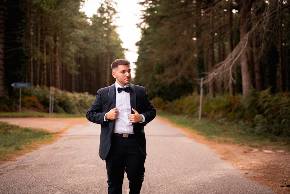 A man in formal attire, including a black tuxedo and bow tie, standing on a gravel pathway in a forested area during daytime. He is adjusting his jacket and looking to his left with a neutral expression. The background shows tall trees with green foliage, a blue directional sign on the left side, and a wooden utility pole with wires on the right. The lighting suggests natural daylight with soft shadows. Although the scene is outside a property, this image could relate to home relocation or furniture transport as part of a broader move process, aligning with house removals services provided by Man with Van Cranham. The setting emphasizes a quiet, natural environment, suitable for visual storytelling about moving logistics or detailed packing and loading routines involving vehicles like vans or trucks.