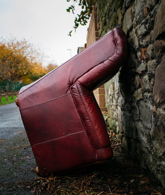 A large, burgundy leather sofa has been placed upright against a stone exterior wall of a building, with one armrest and part of the backrest visible. The sofa is positioned on a paved sidewalk beside a brick wall, with autumn foliage and trees in the background, indicating an outdoor setting. The furniture appears to be in the process of home relocation or being prepared for collection, with no packaging materials or additional items visible. A vehicle, likely a removal van, is not shown but implied by the context of furniture transport services. The scene captures a moment during a house removal or furniture delivery operation carried out by Man with Van Cranham, with the sofa awaiting loading into the van as part of the moving process, illustrating the careful handling of large household items during packing and moving tasks.