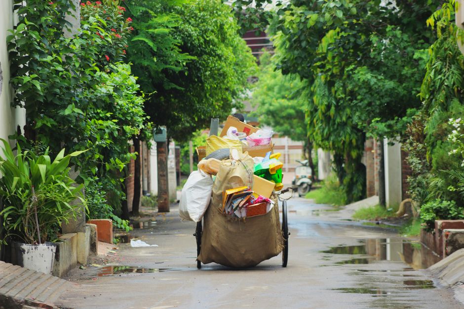 A bicycle parked on a narrow residential street surrounded by green trees and foliage, loaded with large brown and white plastic bags, cardboard boxes, and plastic containers filled with various household items. The items are stacked and secured on the bicycle, some wrapped in plastic or fabric, while others are exposed. The street surface appears wet, with puddles reflecting the greenery and the bicycle. In the background, distant houses and street furniture are visible, with the scene illuminated by soft natural daylight. The image demonstrates the process of collecting bulky waste or recyclable materials, aligning with home relocation or moving services provided by Man with Van Cranham, often involving transportation and logistics of household goods.