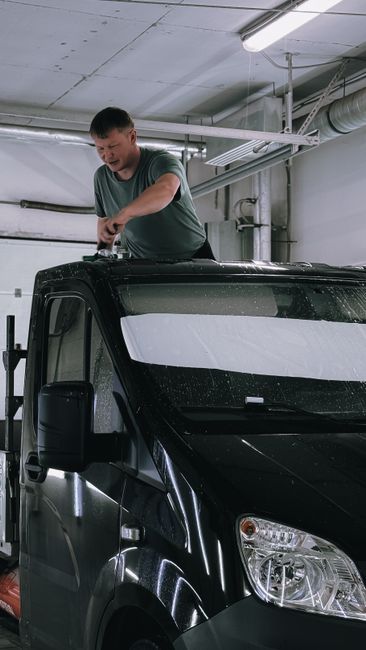 A man with short dark hair, wearing a grey t-shirt, standing inside a parking garage or garage area, is carefully sealing or inspecting a large cardboard box with packing tape. The vehicle, a black van with a white stripe on the front, is positioned nearby with its side mirror visible. The background includes industrial elements such as exposed pipes and ductwork attached to the ceiling, which is illuminated by fluorescent lighting. This scene depicts a home relocation or furniture transport process, with the man preparing items inside the van for moving. The environment appears clean and organized, with the focus on the packing and loading activities typical of professional removals services provided by Man with Van Cranham as part of their house removals offerings.