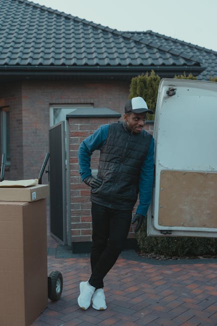 A man wearing a grey cap, blue long-sleeve shirt, and black padded vest stands outside a residential property during daytime, next to an open van used for house removals in Cranham. He has his hands on his hips and appears to be observing the loading process. The van's side door is open, revealing an empty interior prepared for furniture transport or packing materials. In the foreground, there is a cardboard box on a hand trolley, indicating ongoing packing or loading activities. The environment includes a brick pavement, a modern house with a grey tiled roof, a door and window visible on the building, and some greenery, suggesting a typical home relocation scene with professional removals service support by Man with Van Cranham.