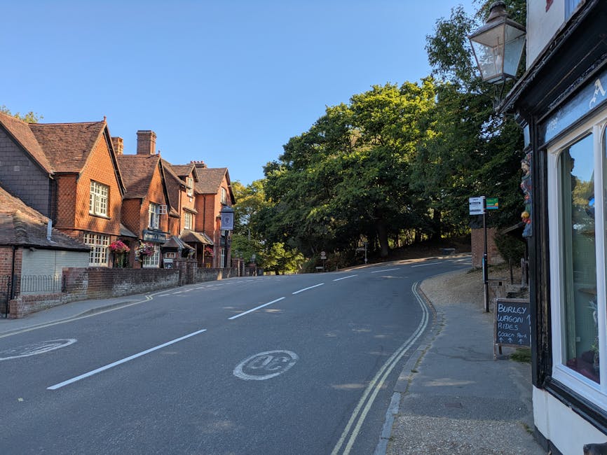 A quiet residential street in Cranham with a slight uphill slope, showing a row of traditional brick houses on the left side, each with small front gardens and decorative window boxes. On the right side, part of a small shop or office with large glass windows and a signboard, situated on the pavement. The roadway is marked with white dashed lines dividing lanes, and there is a 30 miles per hour speed limit painted on the asphalt. A bus stop is visible with a bus stop sign and a timetable on a pole near the pavement. Tall, leafy trees shade the street, creating a pleasant atmosphere suitable for a home relocation or furniture transport process. The scene is captured during daylight with clear blue skies, illustrating the typical environment where professional removals by [COMPANY_NAME] might take place during busy moving days, especially tailored for navigating narrow streets in Cranham.