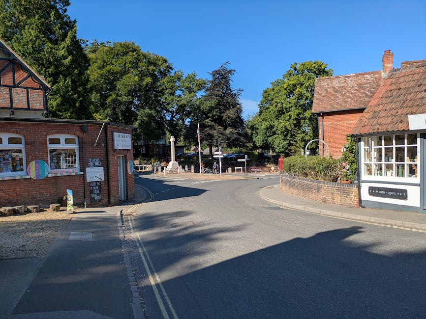 A quiet residential street in Cranham with a narrow, curved asphalt road flanked by brick buildings and green leafy trees under a clear blue sky. On the left, a small brick commercial building with decorative windows and a sign, possibly a café or local shop, has outdoor chalkboard signs and a painted wheel decoration. On the right, a traditional brick house with a tiled roof, white-framed windows, and a small front garden is visible, while behind the buildings, larger trees provide shade and greenery. In the background, a small square or roundabout with a monument or column in the center is visible, surrounded by more trees. The scene, lit by bright daylight, captures a peaceful local area suitable for home relocation or furniture transport, with no visible vehicles or people actively loading or unloading, but the setting reflects a typical environment in which house removals by [COMPANY_NAME] could take place.