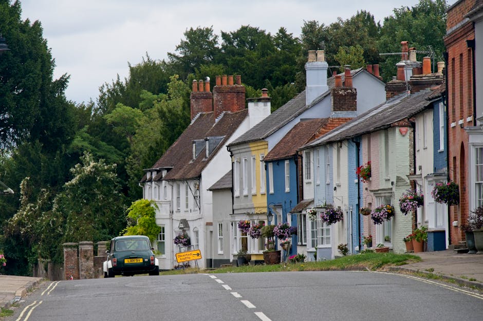 A quiet residential street in Cranham with a slight uphill slope, showing a row of traditional brick houses on the left side, each with small front gardens and decorative window boxes. On the right side, part of a small shop or office with large glass windows and a signboard, situated on the pavement. The roadway is marked with white dashed lines dividing lanes, and there is a 30 miles per hour speed limit painted on the asphalt. A bus stop is visible with a bus stop sign and a timetable on a pole near the pavement. Tall, leafy trees shade the street, creating a pleasant atmosphere suitable for a home relocation or furniture transport process. The scene is captured during daylight with clear blue skies, illustrating the typical environment where professional removals by [COMPANY_NAME] might take place during busy moving days, especially tailored for navigating narrow streets in Cranham.
