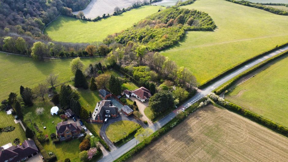 An aerial view of a rural residential property situated amidst green fields and wooded areas, with several houses and gardens. A narrow paved driveway leads from the main road to the front of the hous