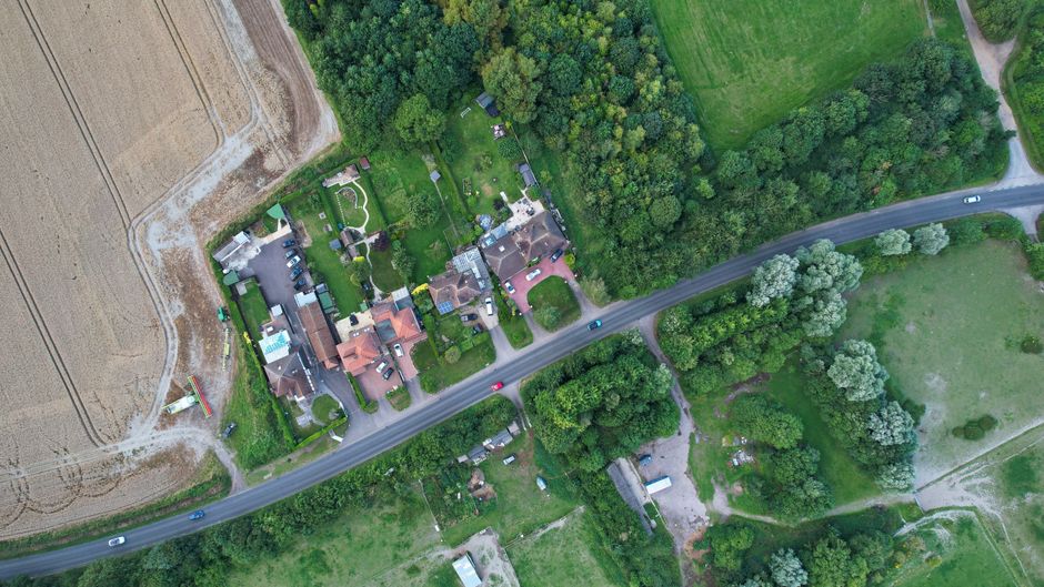 An aerial view of a rural residential property situated amidst green fields and wooded areas, with several houses and gardens. A narrow paved driveway leads from the main road to the front of the hous