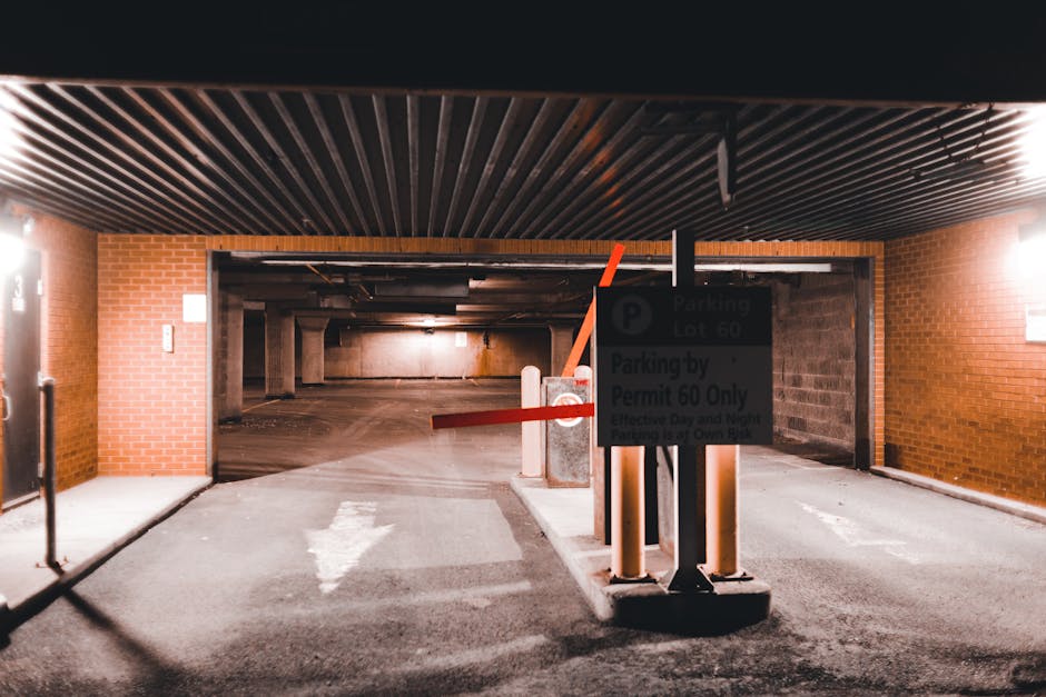 Inside the entrance of a multi-storey parking garage with a dark suspended ceiling featuring parallel black slats, brick walls on both sides, and a concrete ground surface. The area is illuminated by wall-mounted lights on the right side. A parking barrier with red and white striped arms is positioned in front of a sign indicating parking regulations, including restrictions for parking permits and designated parking lot 60. The sign is mounted on black posts anchored in a concrete base, partially blocking the ramp leading into the garage's interior. The ramp curves slightly downward and is flanked by concrete barriers, with visible parking spaces and columns extending into the background. This setting is relevant for house removals, as it depicts a typical vehicle access point for furniture transport and moving logistics, with an emphasis on loading and parking arrangements for professional removals by Man with Van Cranham.