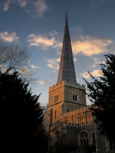 Photograph of a tall, historic church with a pointed spire extending high into the sky, situated in a residential area at sunset. The church features a stone facade with arched windows, decorative battlements along the roofline, and a square tower at the base supporting the slender, tapering spire covered in metallic panels. The sky behind shows a mix of blue and orange hues with scattered clouds, while the foreground includes dark silhouette trees and bushes, framing the building. This image is relevant to house removals and relocation services, illustrating the local architecture where [COMPANY_NAME] might operate, and highlighting the importance of careful planning when moving in and around historic sites.