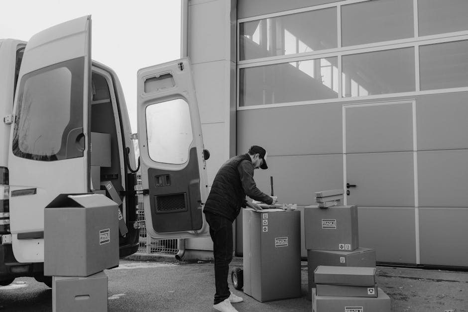 A man wearing a cap and dark clothing is seen standing outside near a large commercial van with its rear door open, engaged in packing or unpacking. He is handling several cardboard boxes, some stacke