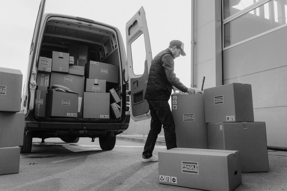 A man wearing a cap and dark clothing is seen standing outside near a large commercial van with its rear door open, engaged in packing or unpacking. He is handling several cardboard boxes, some stacke