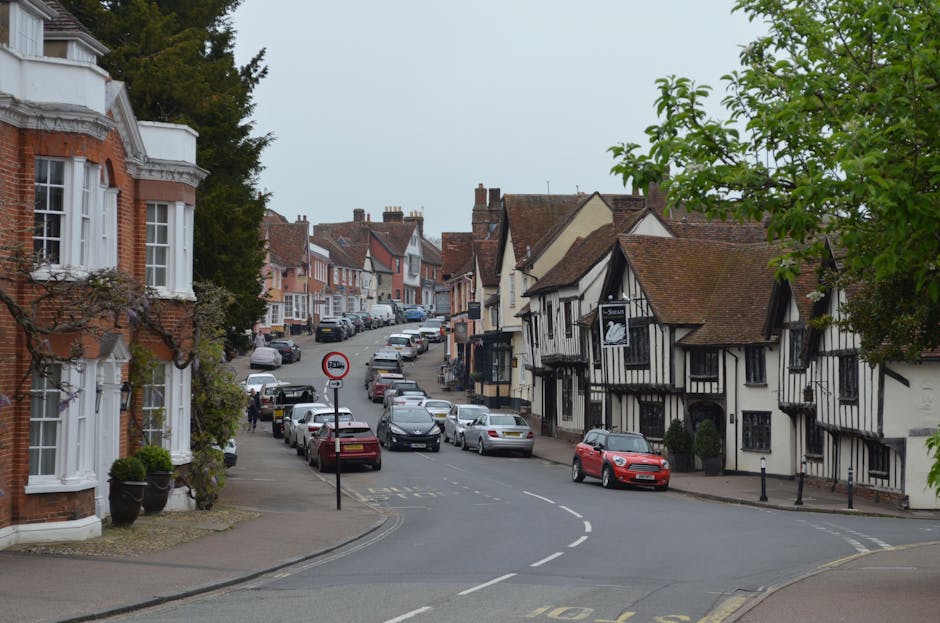 A residential street in Cranham lined with parked cars along both sides, with a combination of traditional and modern houses made of brick and timber, featuring bay windows, pitched roofs, and some with decorative elements. The street slopes slightly uphill, with a number of vehicles including sedans and small vans parked parallel to the curb. In the foreground, a sidewalk runs along the left side of the street, with potted plants near a red-brick house that has white-framed windows. An upright signpost indicating a no-entry zone for certain vehicles is visible in the middle ground. On the right side, greenery from a tree partially frames the scene, with overcast weather providing diffuse natural lighting. This environment illustrates an urban neighbourhood setting suitable for home relocation activities and furniture transport, aligning with professional removal services such as those offered by Man with Van Cranham, who conduct packing, loading, and moving logistics within residential areas.