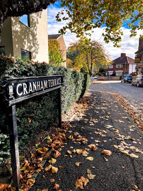 A quiet street scene in Cranham featuring a small brick building on the left with decorative window displays, and a larger brick house with a sloped tiled roof on the right, both surrounded by tall green trees under a clear blue sky. In the foreground, a paved road curves gently to the left, with a double yellow line along the curb. A white van from Man with Van Cranham is parked partly on the pavement, with the back of the vehicle extending into the road, indicating the loading process for a home relocation. The vehicle's sliding side door is open, and furniture, boxes wrapped in protective plastic and cardboard, and packing materials are visible being loaded onto or off the van. Equipment such as trolleys and straps are likely being used to facilitate the furniture transport. This scene captures the process of packing and loading household items for a same-day removal service in Cranham, highlighting the logistical aspect of house removals and professional moving services in a residential area.
