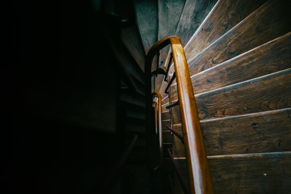 A view looking down a narrow staircase with wooden steps and a polished wooden handrail, illuminated by natural light from an unseen window or doorway. The stairs are inside a residential property, with the surrounding wall partially visible in dark shadows. The staircase appears to be part of a home with traditional wooden flooring, and the scene captures a moment during the house move process, potentially involving furniture transport or packing and moving activities. Man with Van Cranham, a professional removals company, specializes in house relocation services including navigating tight staircases in Cranham flats, ensuring safe and efficient removal solutions for clients. The photo emphasizes the staircase's confined space, highlighting the importance of careful planning and equipment such as blankets or straps during the moving process.