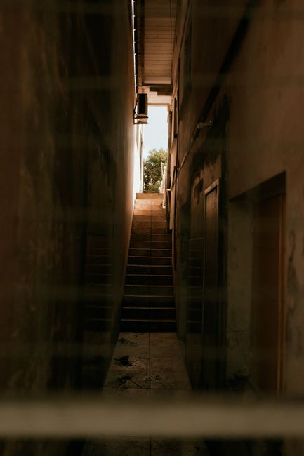 A narrow indoor staircase with wooden steps and a metal railing, ascending between two walls in a residential property. The walls are plain and beige, with some marks and slight discoloration. At the top of the staircase, sunlight illuminates the area, revealing a small outdoor space with trees and greenery visible through an open door or window. The staircase appears to be part of a compact flat or apartment, often found in Cranham, and is accessed during home relocation or moving processes. The image captures the confined space typical of stairs in older buildings, which can pose challenges for furniture transport and packing during removals. Man with Van Cranham, a professional removal company, likely involved in such a home relocation, could be coordinating safe and efficient strategies to navigate tight staircases like this during a furniture transport or packing and moving operation, utilizing equipment such as trolleys or blankets to protect belongings and ensure safety.