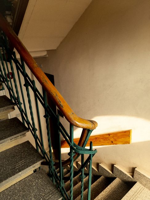 A view looking down a narrow staircase with wooden steps and a polished wooden handrail, illuminated by natural light from an unseen window or doorway. The stairs are inside a residential property, with the surrounding wall partially visible in dark shadows. The staircase appears to be part of a home with traditional wooden flooring, and the scene captures a moment during the house move process, potentially involving furniture transport or packing and moving activities. Man with Van Cranham, a professional removals company, specializes in house relocation services including navigating tight staircases in Cranham flats, ensuring safe and efficient removal solutions for clients. The photo emphasizes the staircase's confined space, highlighting the importance of careful planning and equipment such as blankets or straps during the moving process.