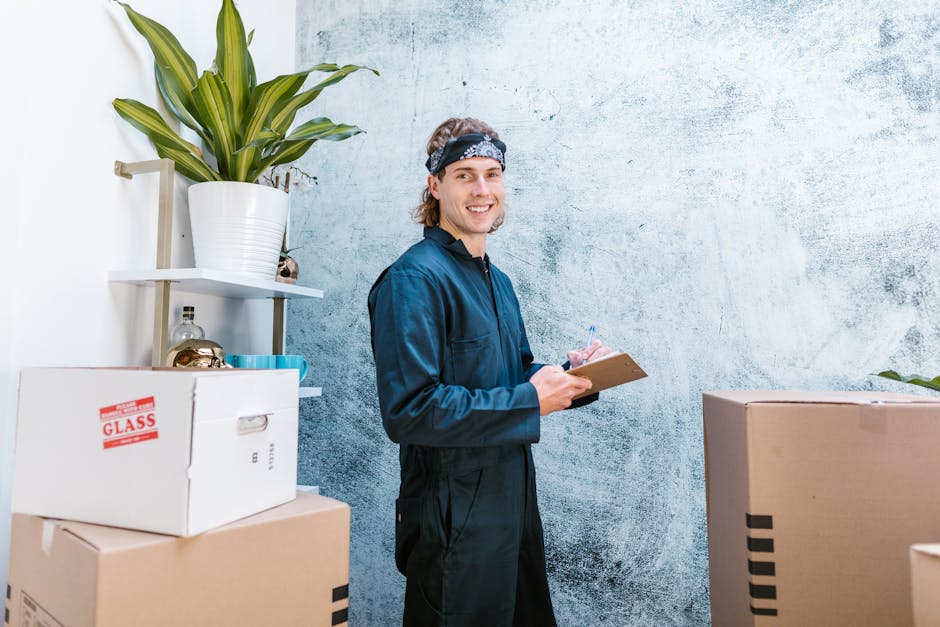 A man with long wavy hair, wearing a dark blue work jumpsuit and a patterned headband, stands indoors next to several cardboard boxes, one labeled 'GLASS' in red, and a white shelving unit holding a large potted plant and other small items. The scene appears to be part of a home relocation process, with the man possibly inspecting or organizing packing materials during furniture transport or packing and moving activities. The background features a textured light blue wall with a slightly weathered appearance, and the lighting is bright and natural, highlighting the scene's clarity. This image is associated with house removals and emergency relocation services offered by Man with Van Cranham.