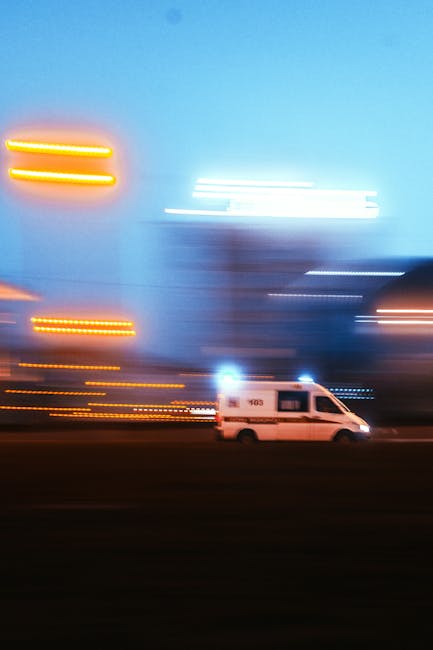 A white van from Man with Van Cranham parked on a city street during dusk or evening with a blurred background of illuminated buildings and streetlights, captured in motion. The van is situated on a paved area just beside the road, with no visible loading equipment or furniture around it. The background features tall buildings with bright, elongated light streaks due to motion blur, and the sky is a gradient of blue and darker tones. The scene illustrates a busy urban environment where home relocation or furniture transport services by Man with Van Cranham could take place, especially during an urgent move or emergency removal, highlighting the logistical aspect of moving and transportation in a city setting.