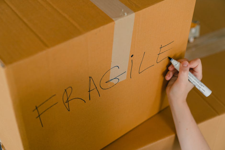 A person's hand holding a black marker pen is writing the word 'FRAGILE' in uppercase letters on a cardboard moving box. The box is light brown and appears to be part of a larger stack, with other similar boxes visible beneath it. The writing is centered on the front face of the box, which is taped with clear packing tape along the top edge. The background shows a wooden floor and possibly part of a home interior, suggesting an indoor setting during the packing phase of a home relocation. The image emphasizes careful handling and packing practices, typical in professional house removals and furniture transport services provided by Man with Van Cranham, illustrating the importance of clearly labeling fragile items during packing and moving.